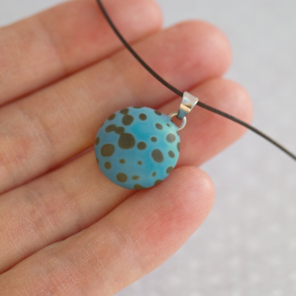 Rankų darbo pakabukas su tamsiais taškeliais žaliai mėlynos spalvos fone 