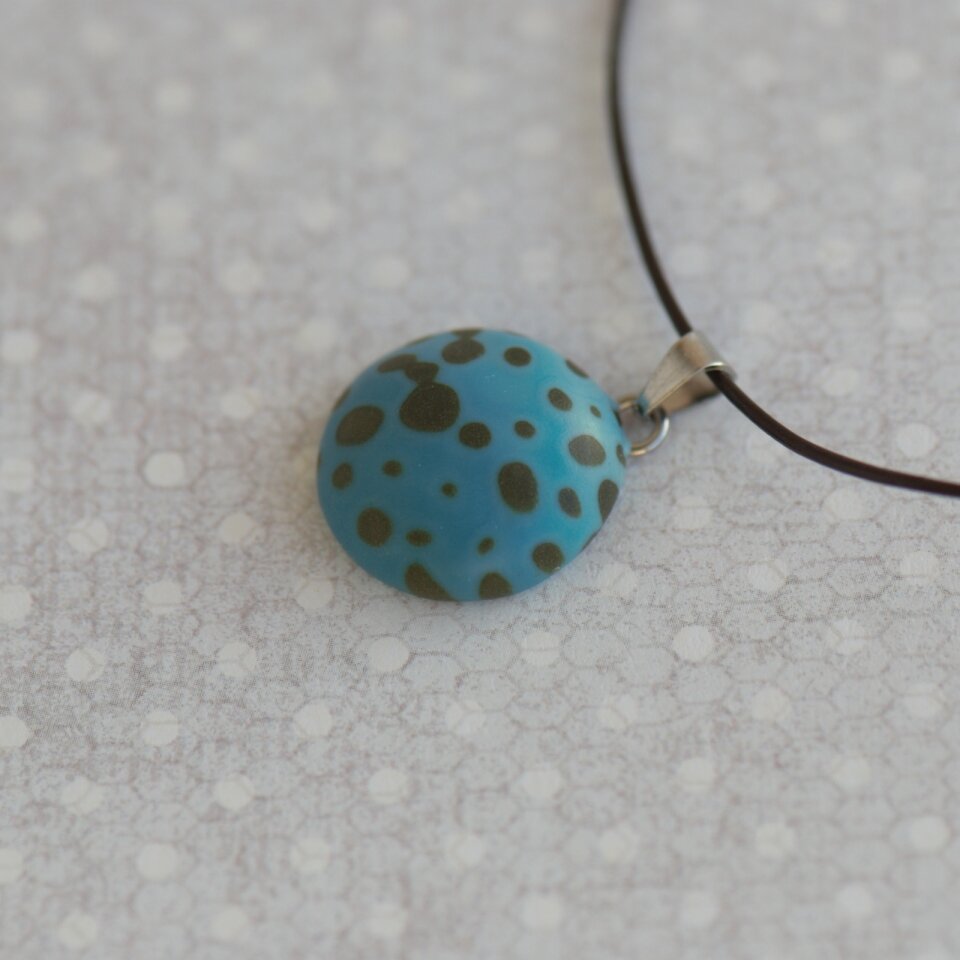 Rankų darbo pakabukas su tamsiais taškeliais žaliai mėlynos spalvos fone 